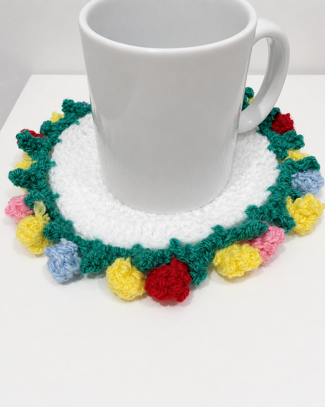 A white ceramic mug placed on a crocheted circular coaster with a green border and colorful pom-pom accents in yellow, pink, blue, and red, set on a white surface.