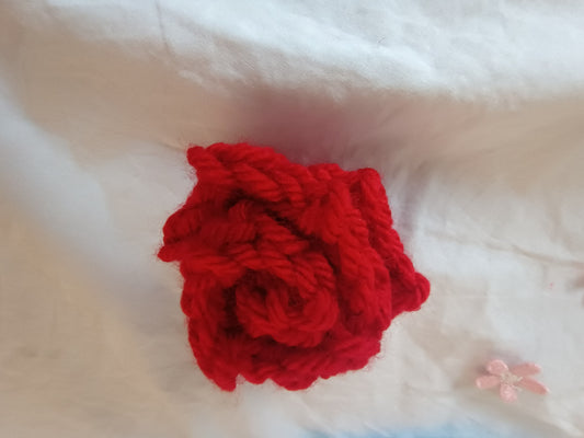 A bright red crocheted yarn rose accessory, likely a hair clip or hair pin, displayed on a soft white fabric background.
