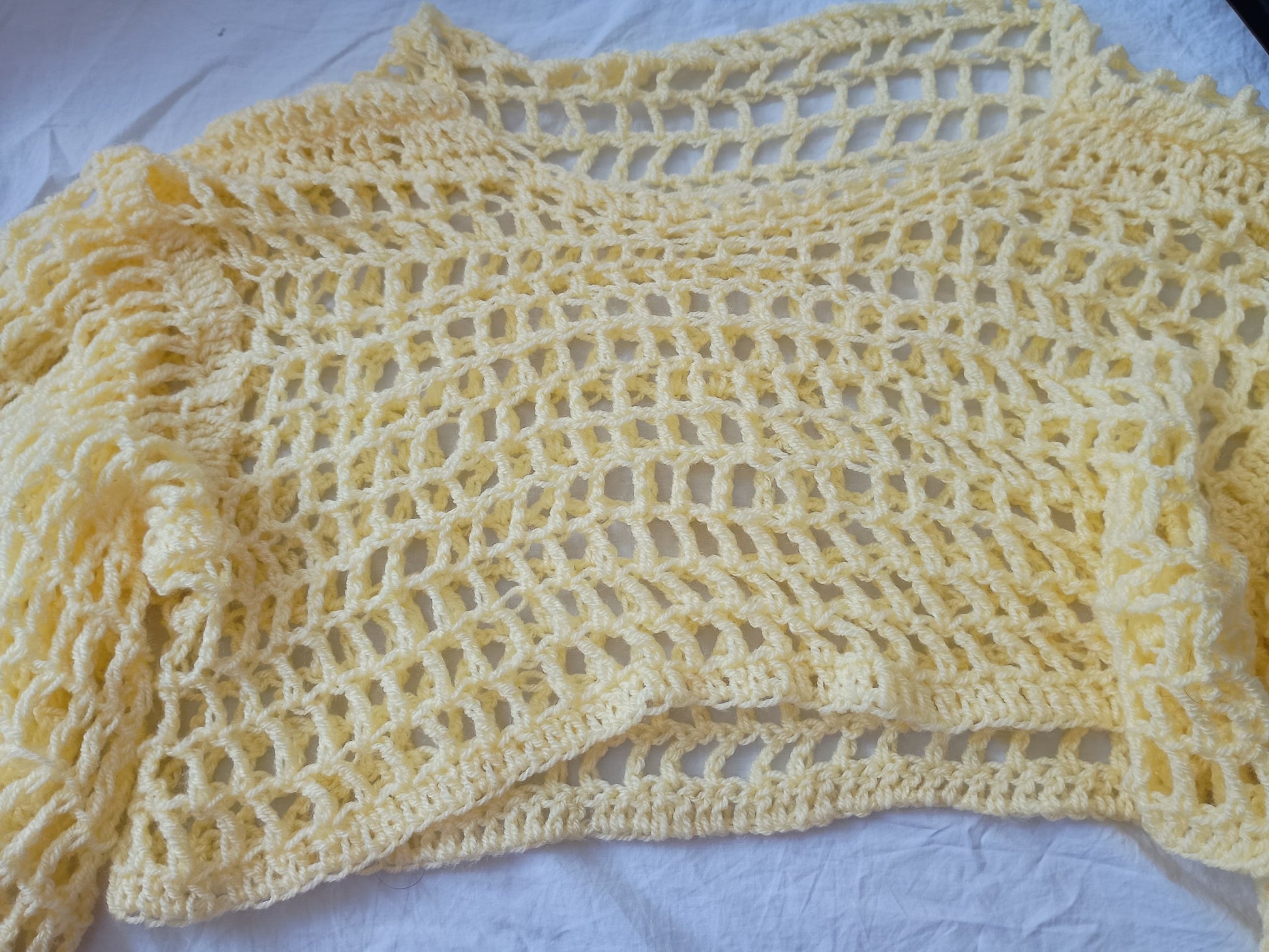 A pale yellow crochet shrug or open-knit cardigan with long sleeves, laid flat on a white fabric background.